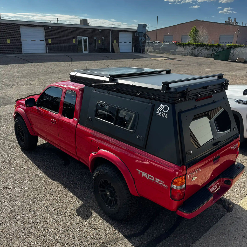 A red Toyota Tacoma pickup truck parked outdoors on asphalt, fitted with a large black hardtop canopy over the bed and a black roof rack system. The canopy has side windows and a rear access door, with a visible "Madix Outdoors" logo on the side.