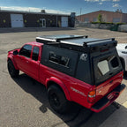 A red Toyota Tacoma pickup truck parked outdoors on asphalt, fitted with a large black hardtop canopy over the bed and a black roof rack system. The canopy has side windows and a rear access door, with a visible "Madix Outdoors" logo on the side.