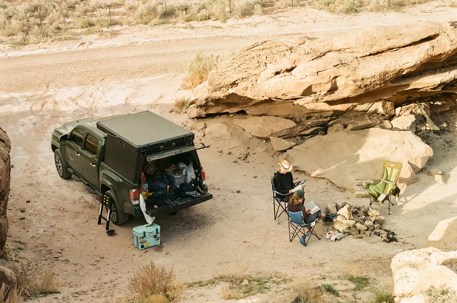 Green Toyota Tacoma with truck bed camper in the desert with someone sitting by a campfire.