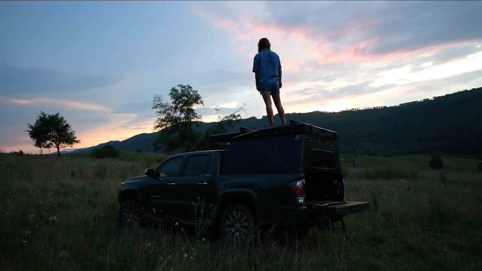 Person standing on a truck bed camper in a field at sunset